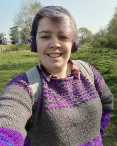A white woman wearing a grey and purple colour block sweater and headphones standing in a field of grass, smiling