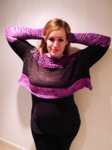 A white woman wearing a black and pink colour-blocked knit sweater with her arms behind her head against a cream background.